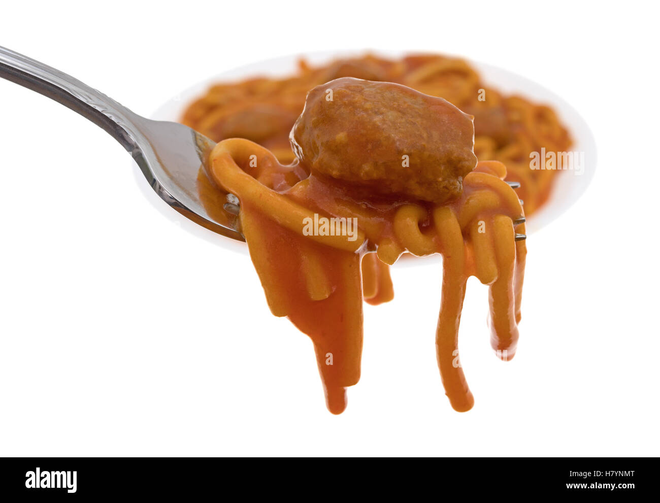 Side view of a meatball atop spaghetti on a fork in the foreground with a plate of food in the background. Stock Photo