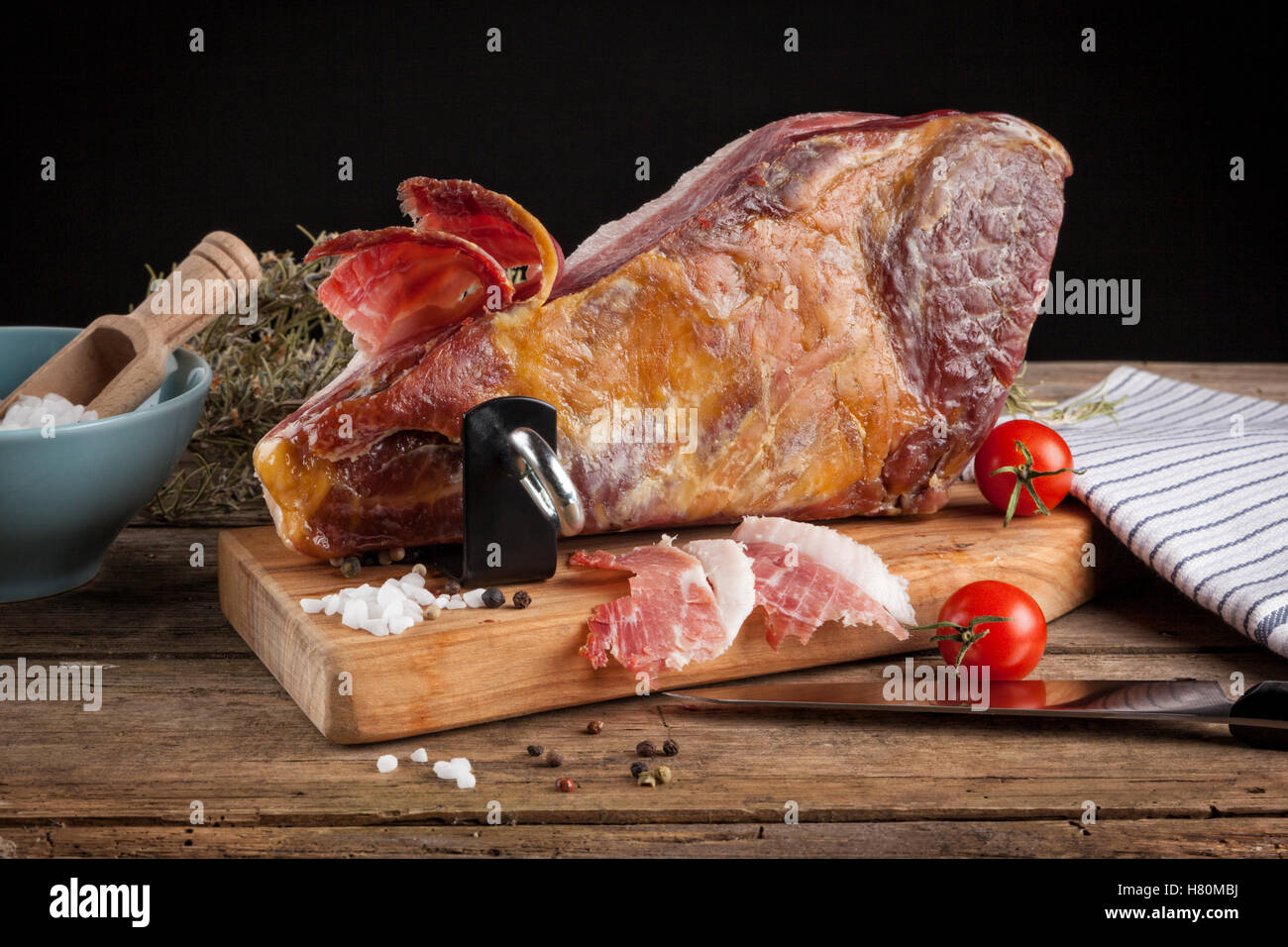 Still-life with dry-cured Serrano ham on ham holder, chopping knife, salt, pepper, tomatoes on rustic wooden table Stock Photo