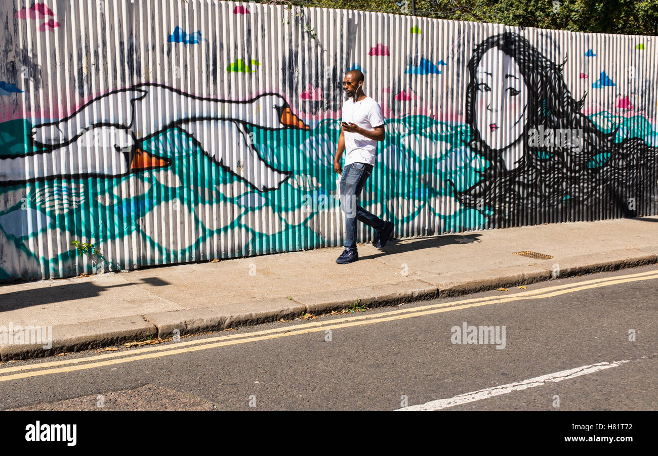 Young black man wearing sunglasses walking in front of a wall with graffiti representing two swans flying and the face of woman Stock Photo