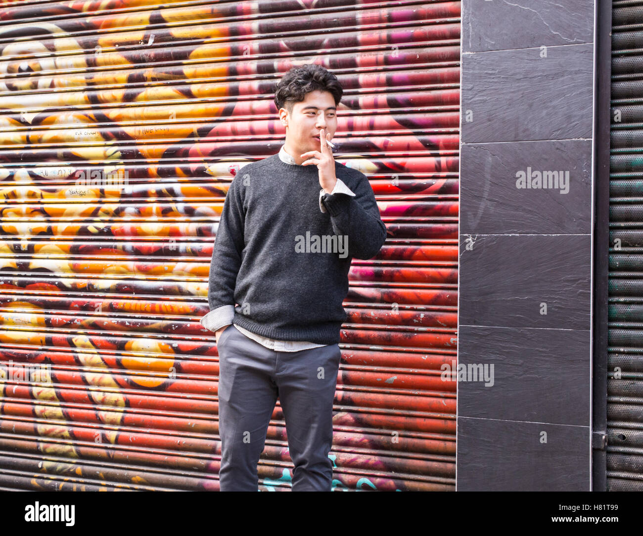 Young Asian man smoking a cigarette in Brick Lane in front of a metal shutter covered in graffiti in the cool area of Shoreditch Stock Photo