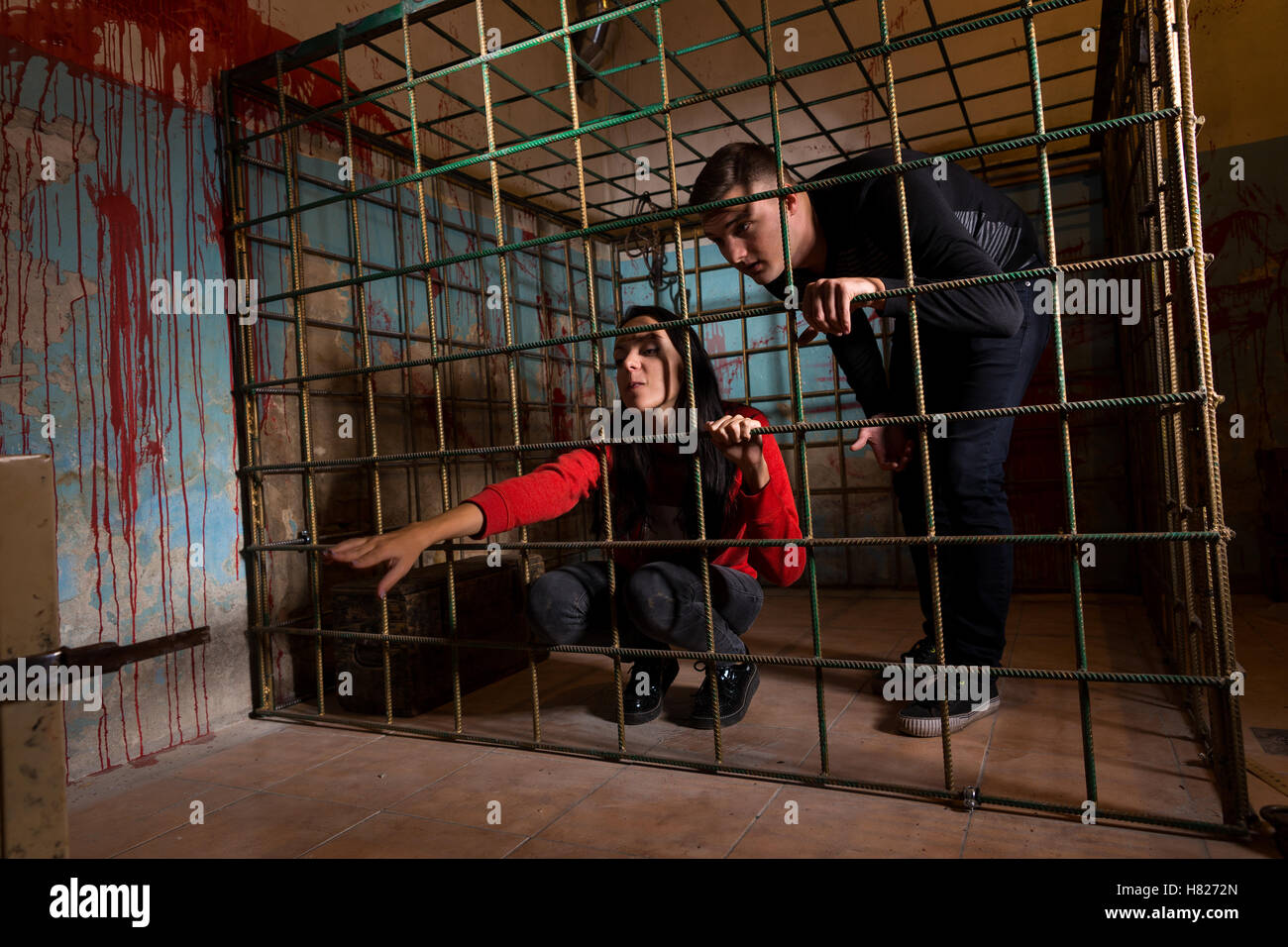 Two victims imprisoned in a metal cage with a blood splattered wall behind them, girl pulling her hand through the bars and tryi Stock Photo