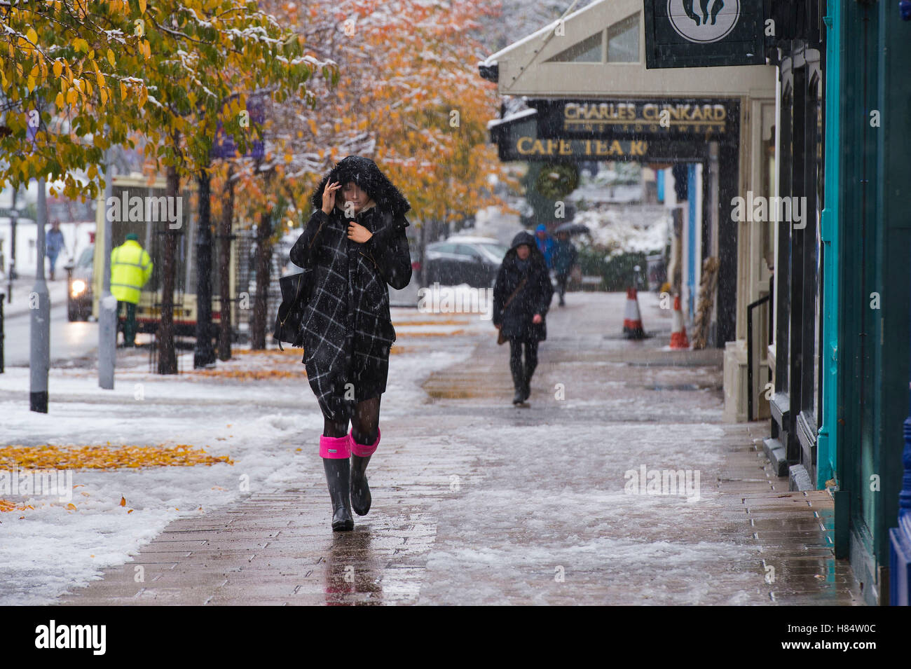 Ilkley, West Yorkshire, UK. 9th November 2016. It is snowing and pedestrians (wearing wellington boots, scarves and winter coats with hoods up) are walking past shops on The Grove. A young woman (hood over her head,  scarf, wellies) is in the foreground. Ilkley's first snowfall of 2016. Credit:  Ian Lamond/Alamy Live News Stock Photo