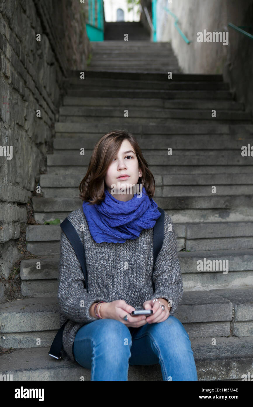 Location shot of a lonely sad teen girl wearing grey sweater and blue scarf sitting on steps with her backpack Stock Photo