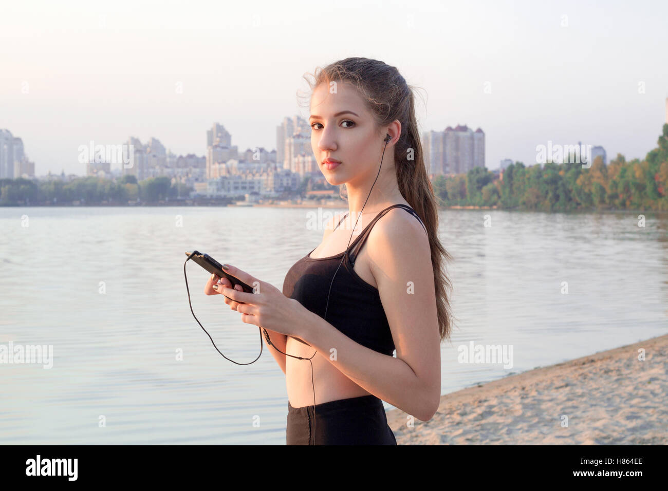 Young pretty slim fitness woman stands at river coast in the morning listening to the music via mobile cell phone player earphon Stock Photo
