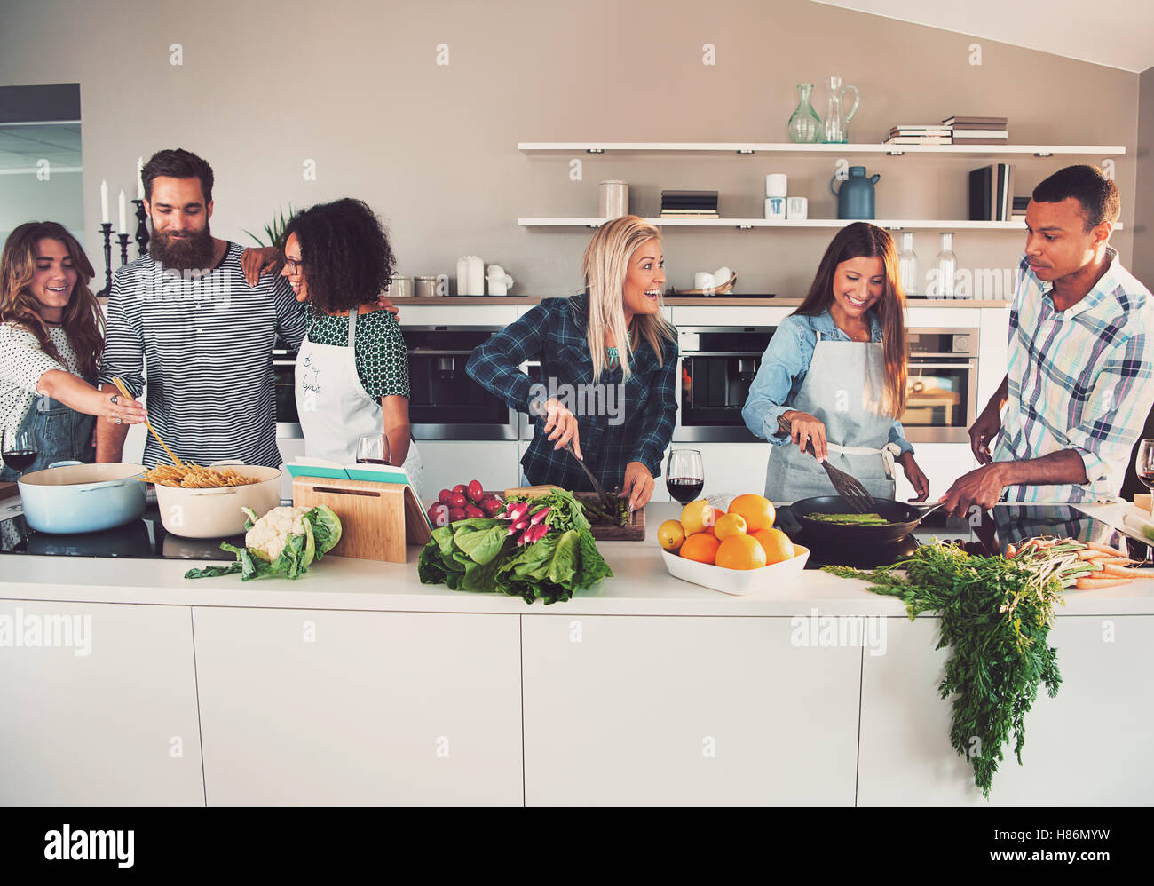 Six mixed Black and white friends preparing vegetables and pasta at long table in kitchen Stock Photo