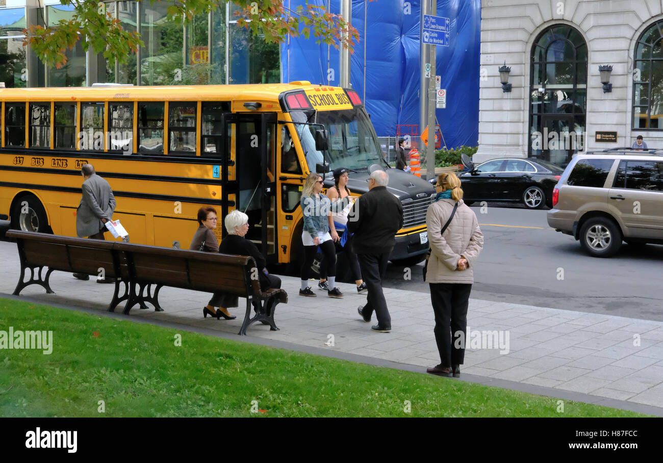 North American styled school bus seen parked on a side road in a busy city, next to a public park, with the public walking past. Stock Photo