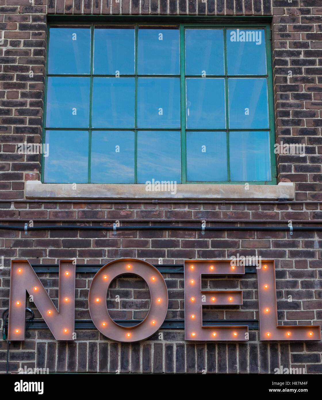 A lit Noel sign made of metal hangs on a brown brick wall. A divided light window above reflects a blue sky. Stock Photo