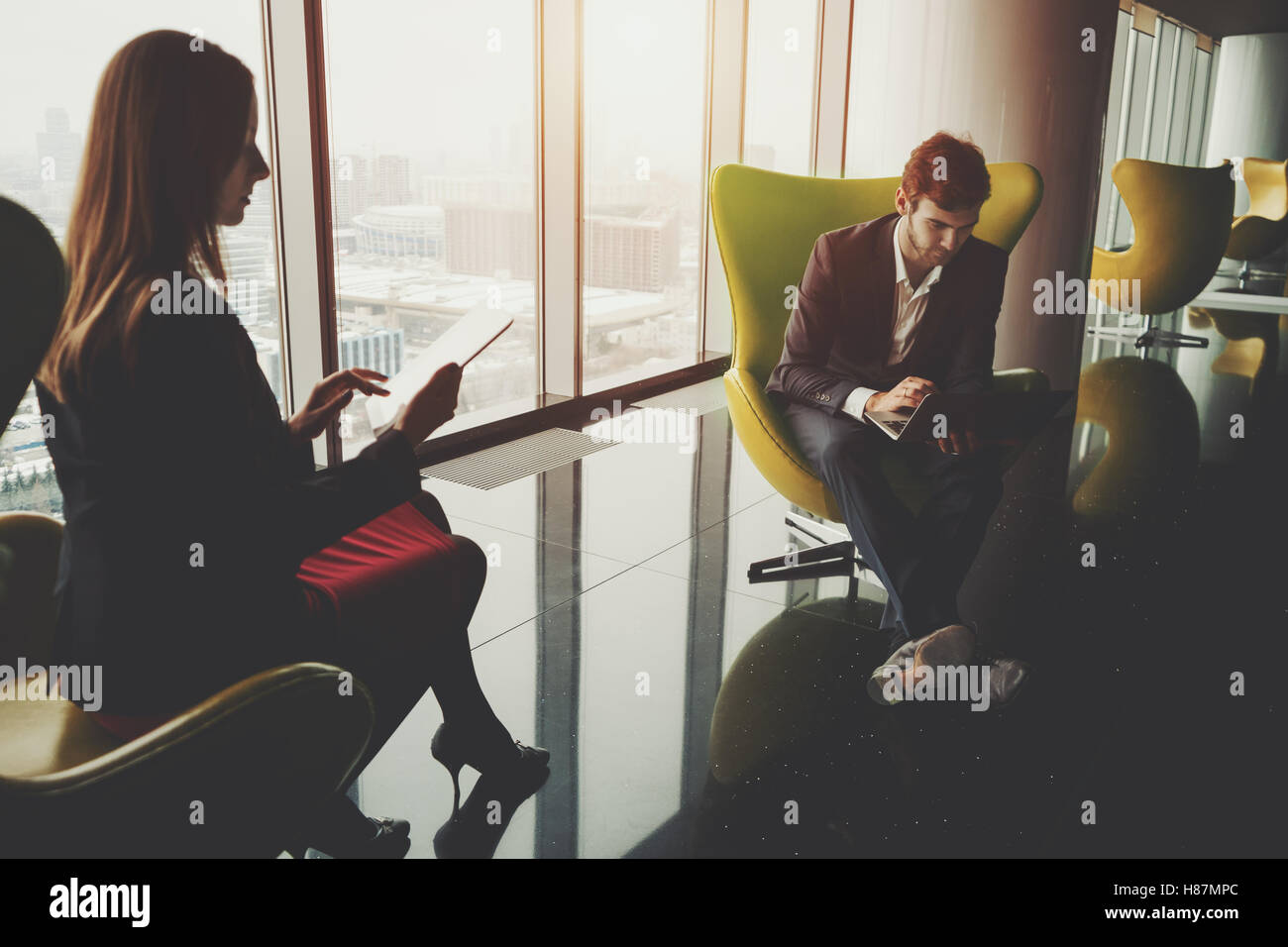 Businessman in formal suite with a laptop and businesswoman in red dress and jacket sitting in front of each other on yellow arm Stock Photo
