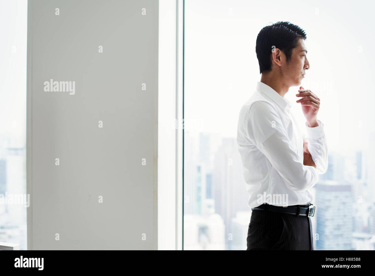 A businessman in the office standing by a large window with arms folded, looking over the city. Stock Photo