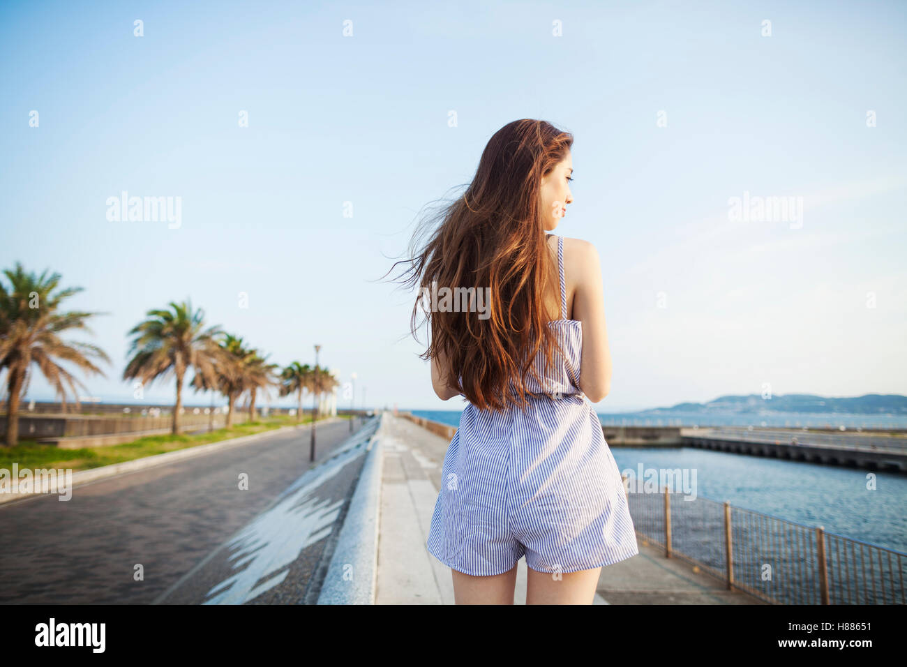 Back view of young woman with long red hair standing in open space by a road on the coast. Stock Photo