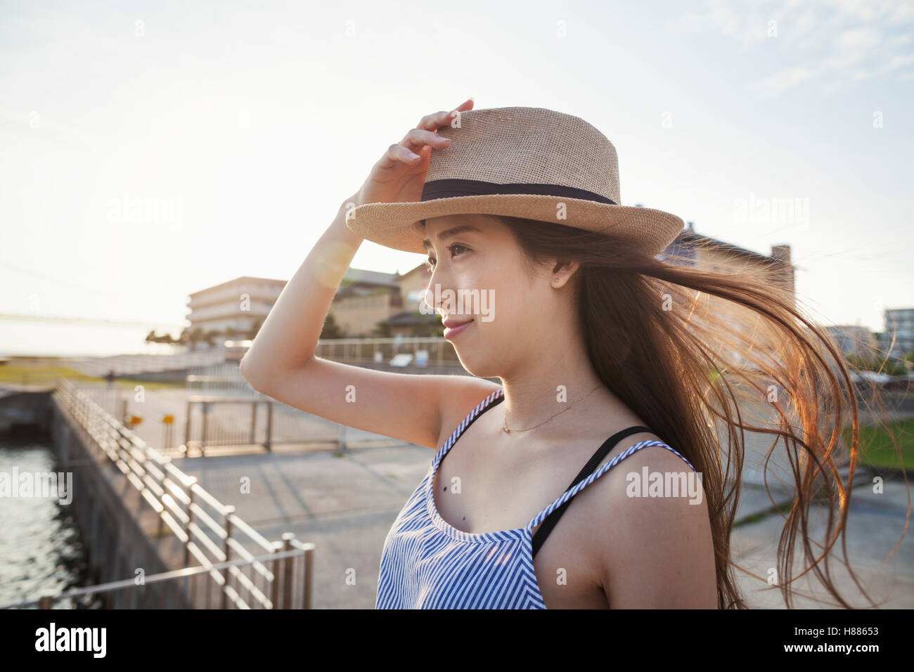 Young woman standing on a pier by water holding her straw hat on her head. Stock Photo