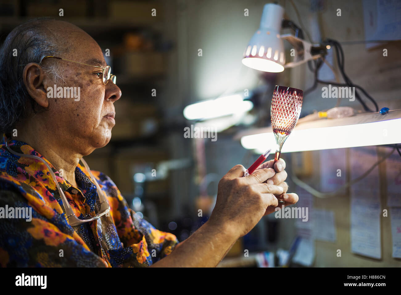 glass maker's studio workshop, man inspecting red wine glass with cut glass decoration against the light. Stock Photo