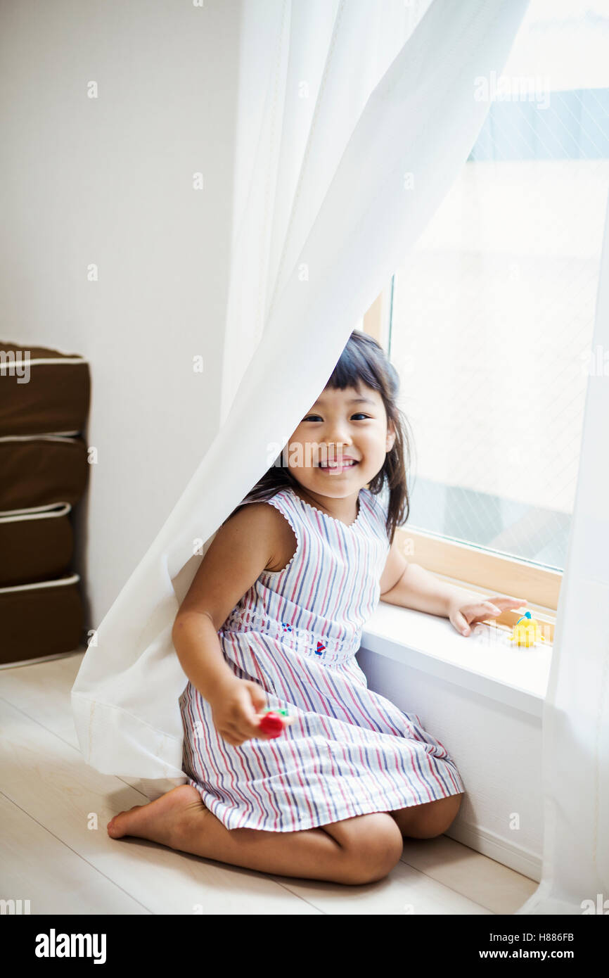 Family home. A girl playing by a window, hiding behind the net curtain. Stock Photo