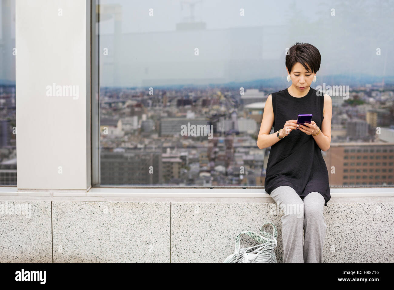 A woman seated by a window with a view over a large city with her back to the view, using her smart phone. Stock Photo