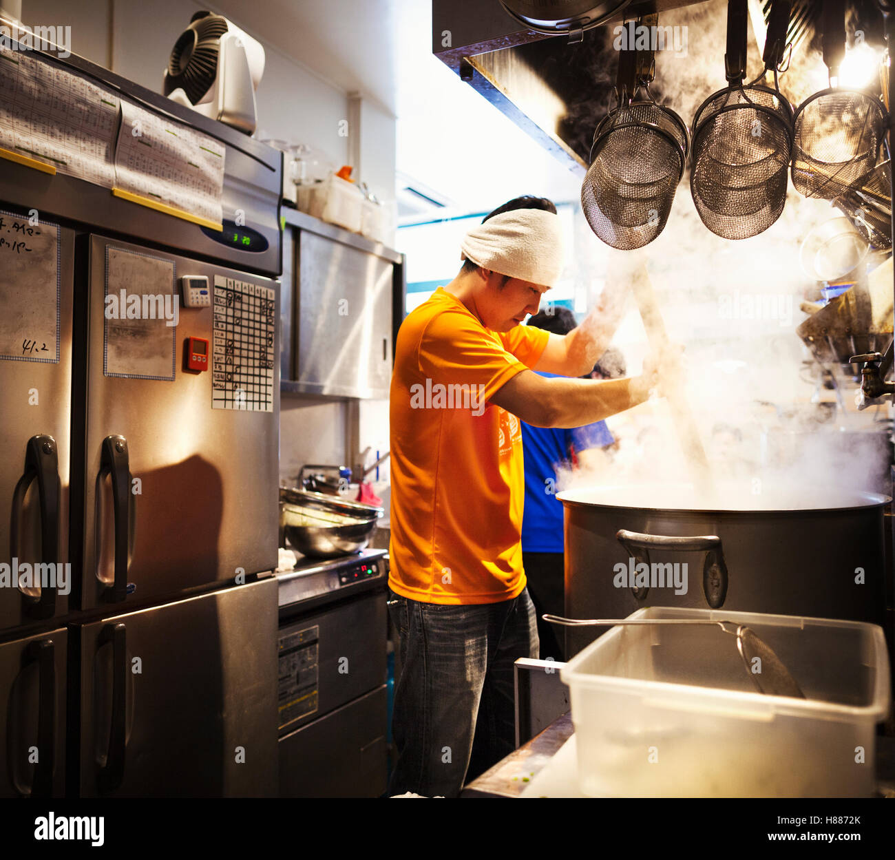 The ramen noodle shop, staff preparing food. Two men stirring a vat of noodles. Steam rising, heat, cooking, bandana small kitchen Stock Photo