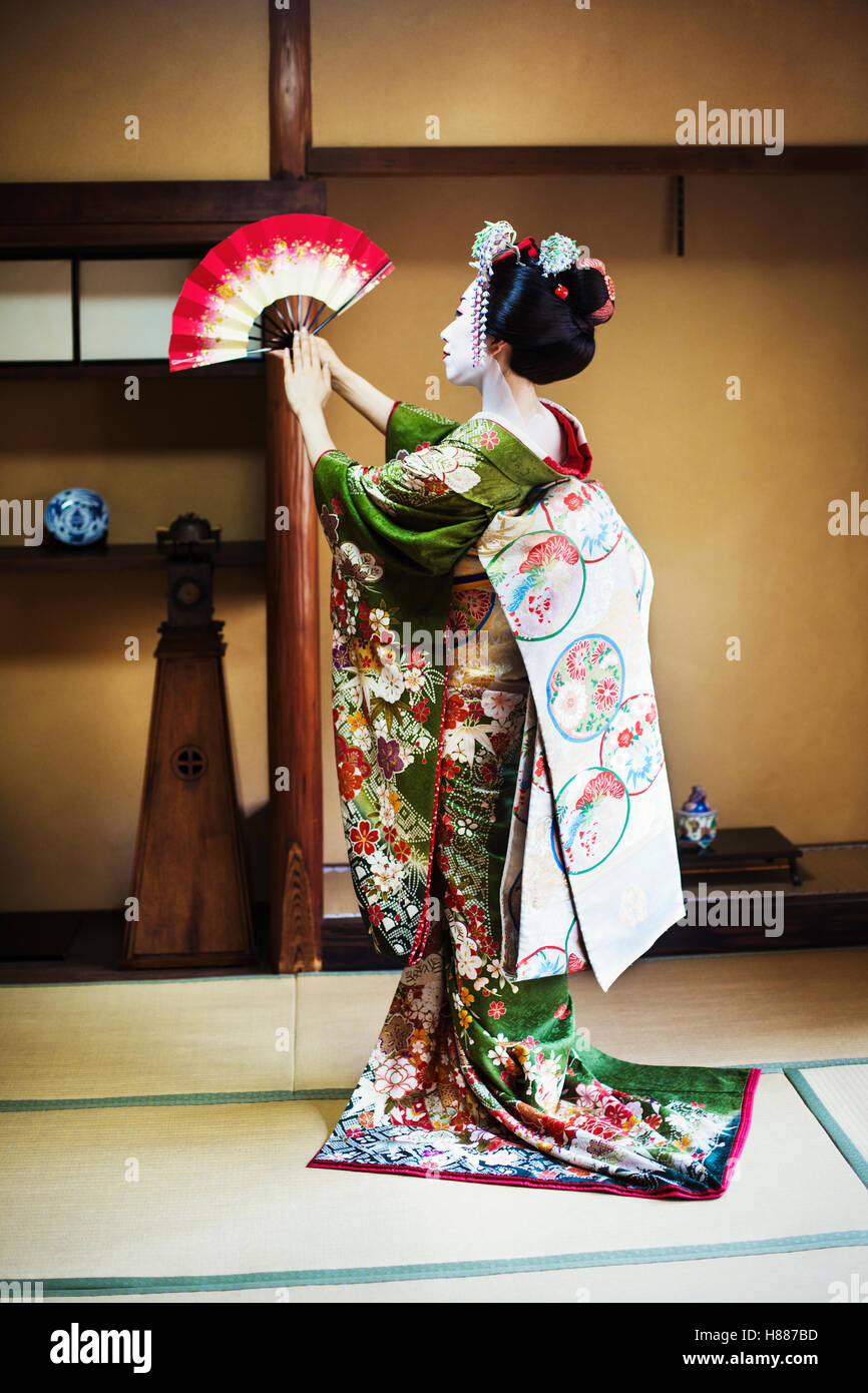 A woman dressed in the traditional geisha style, wearing a kimono and obi, Standing in a classic pose with fan raised Stock Photo