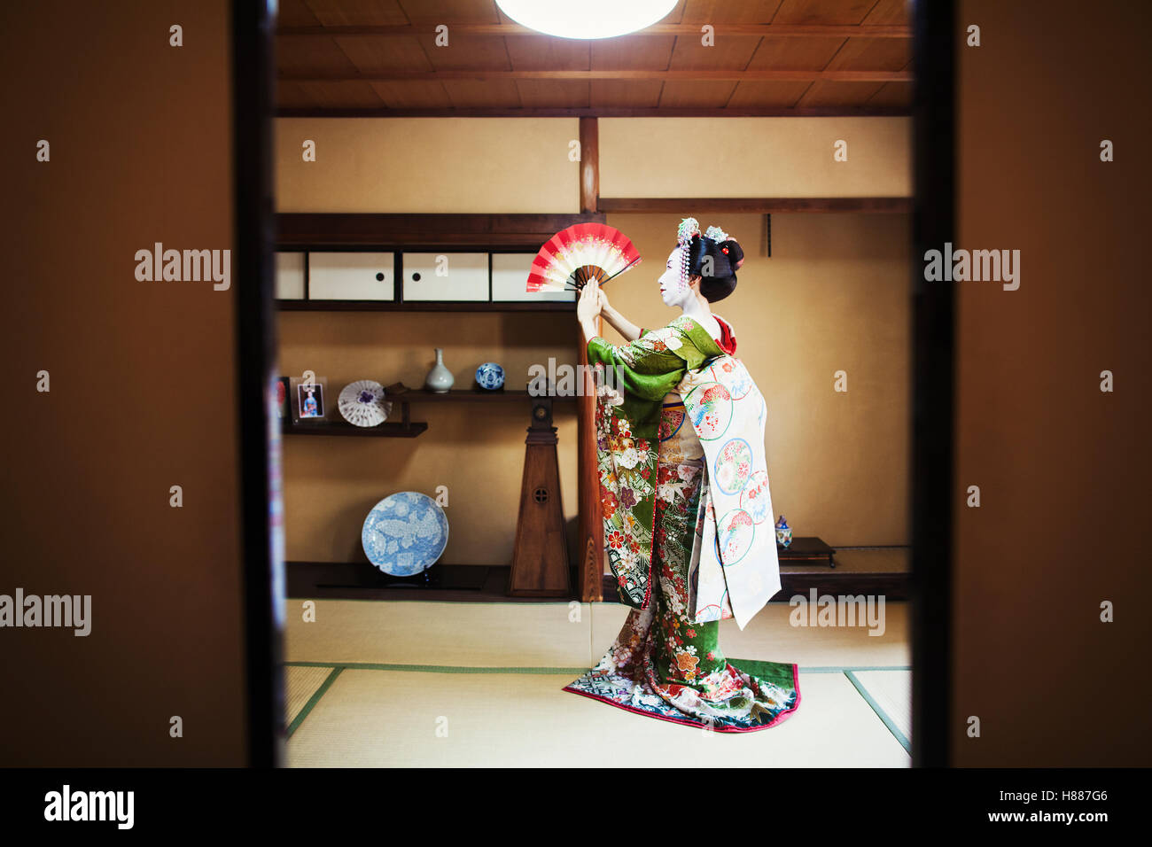 A woman dressed in the traditional geisha style, holding an open fan up, her kimono arranged on the floor. Stock Photo