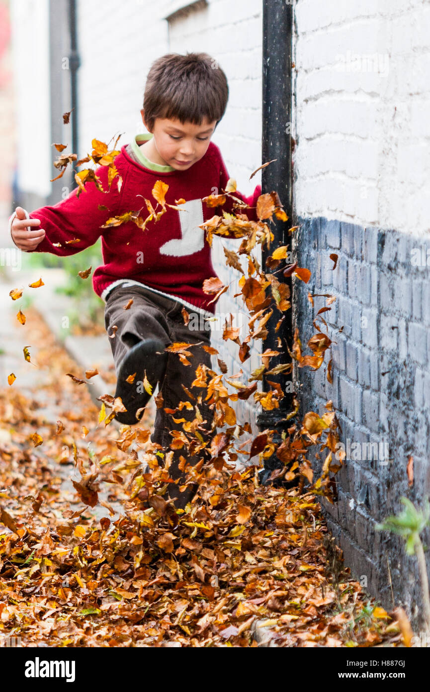 England, Sandwich. Young Caucasian boy, 7-9 years old, in red jumper, kicking leaves up in narrow village street. Directly facing viewer. Autumn. Stock Photo