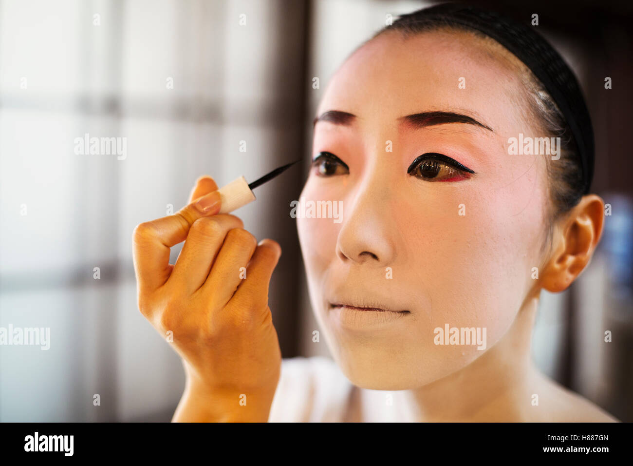 Geisha woman with traditional white face makeup painting on heavy eyeliner with a brush. Stock Photo