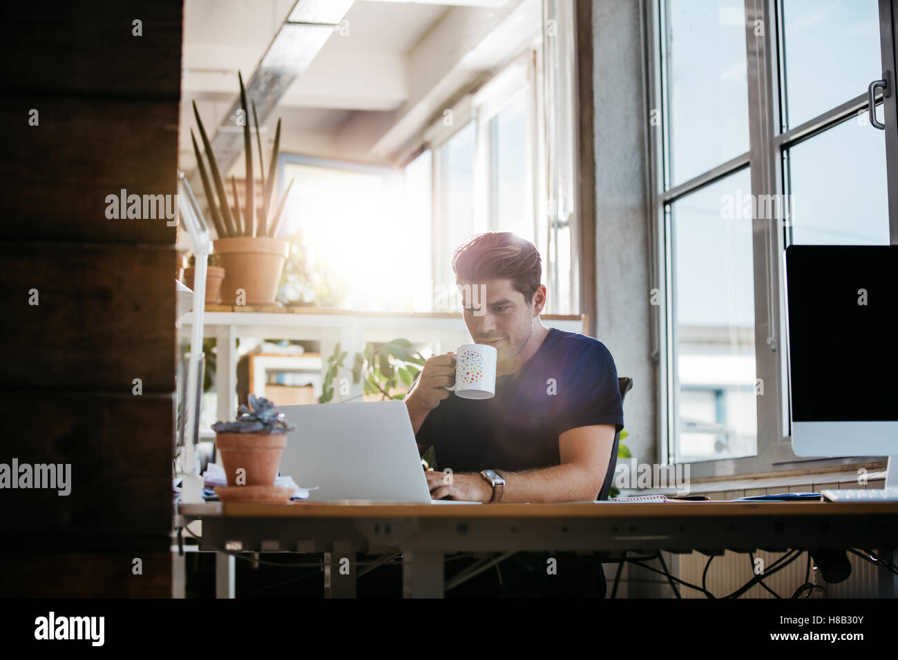Relaxed young man sitting at his desk working on laptop and drinking coffee. Business man in modern workplace. Stock Photo