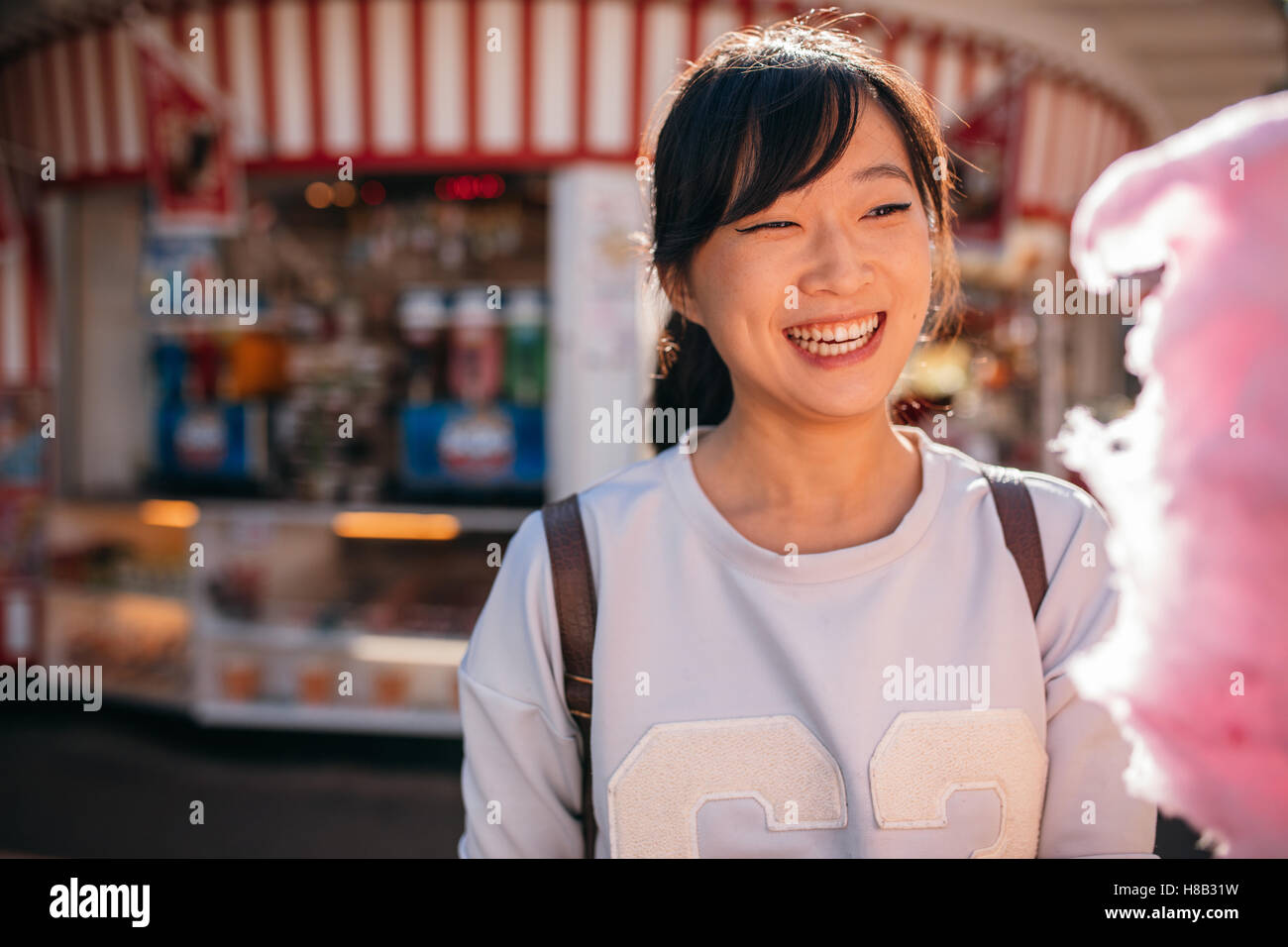 Shot of young asian woman at amusement park with cotton candy floss. Smiling female with candyfloss at fairground. Stock Photo