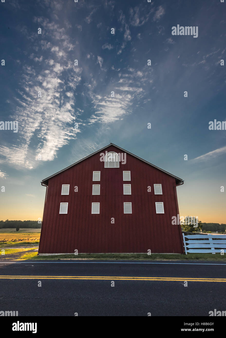 Cloudy Sunset Sky Above Red Barn Towering Over Road Stock Photo