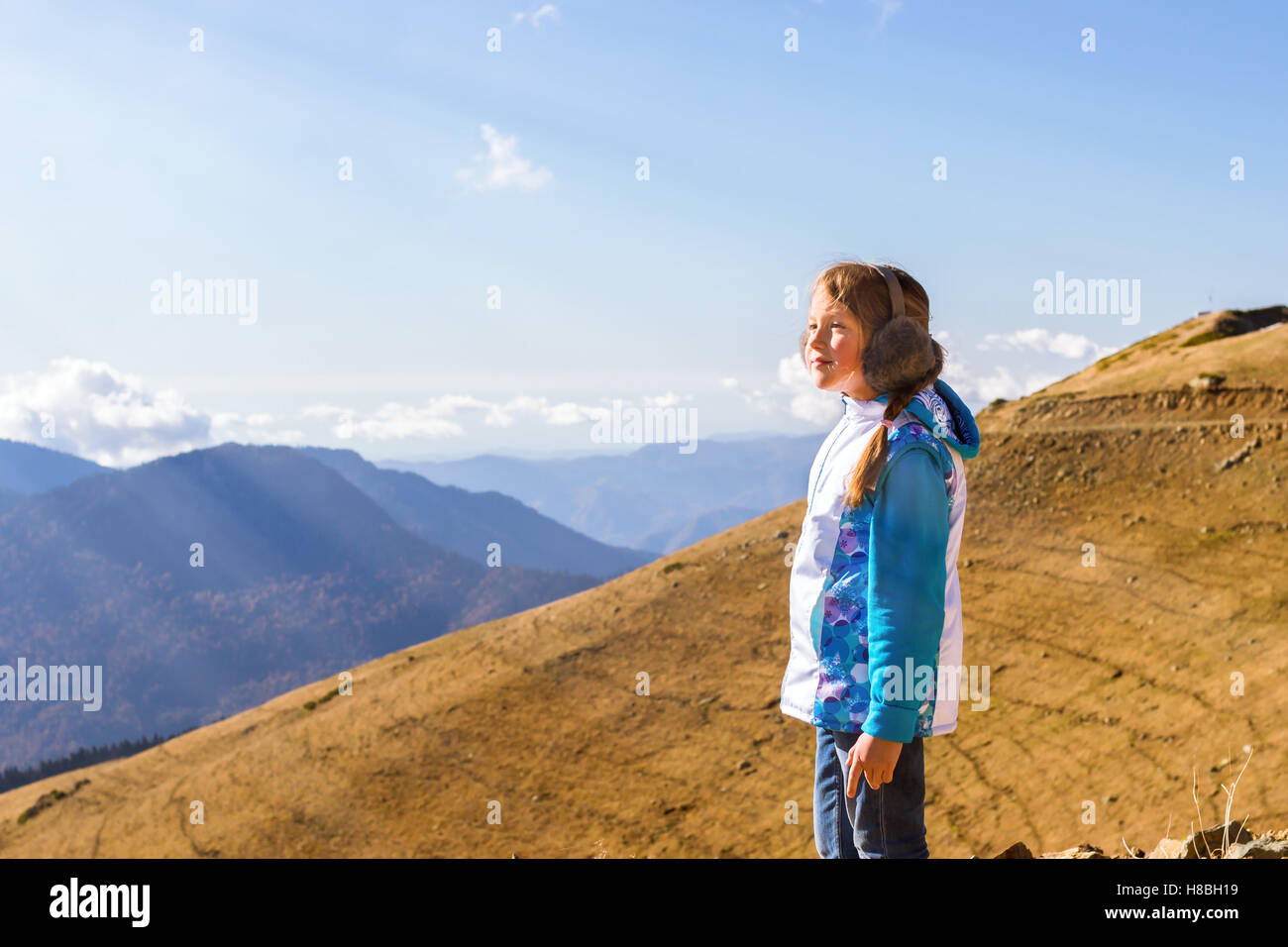 Girl with a pigtail, dressed in sports jacket and warm fluffy headphones, admires of autumn mountain landscape, Sochi, Russia Stock Photo