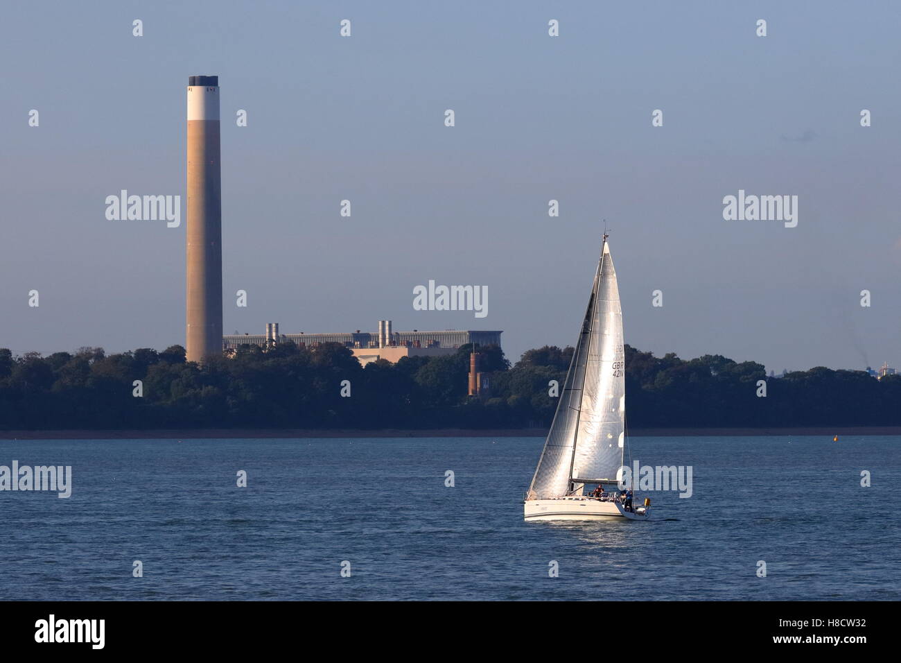 Silver sailed yacht in evening sunshine on The Solent balanced against Fawley power station chimney on Southampton Water Stock Photo