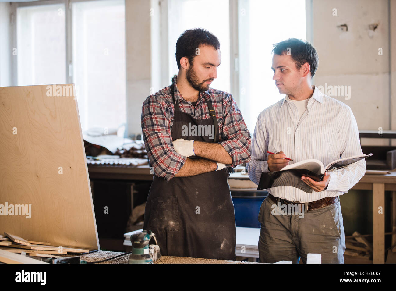 Happy male carpenter showing something to coworker at his notebook papers in workshop Stock Photo