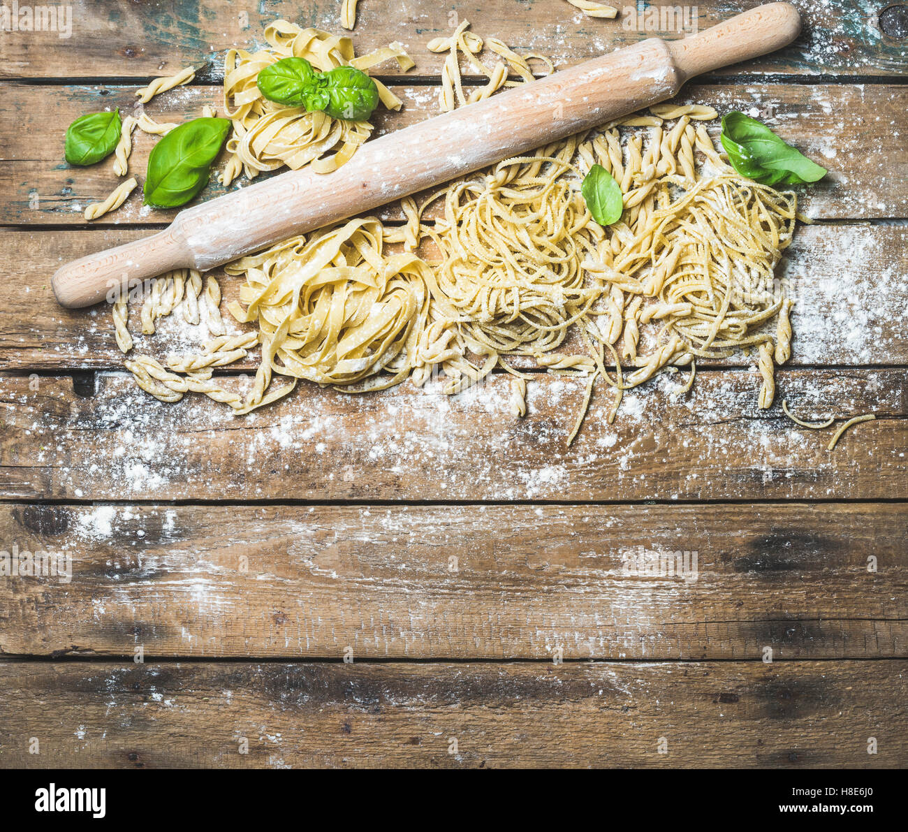 Various homemade fresh uncooked Italian pasta with flour, green basil leaves and plunger on shabby rustic wooden background, top Stock Photo