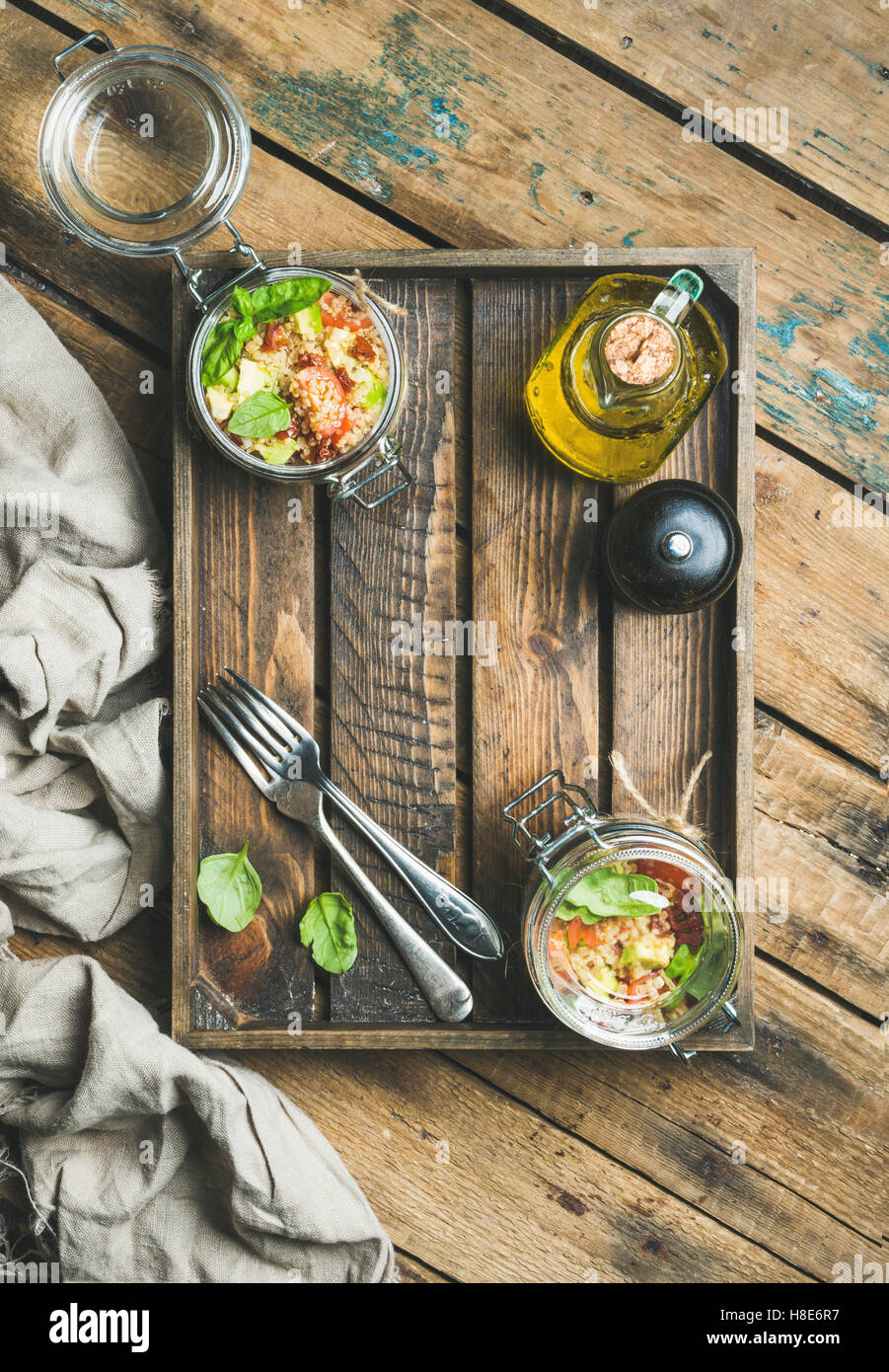 Healthy glass jar quinoa salad with cherry and sun-dried tomatoes, avocado, basil in wooden box over rustic background, top view Stock Photo