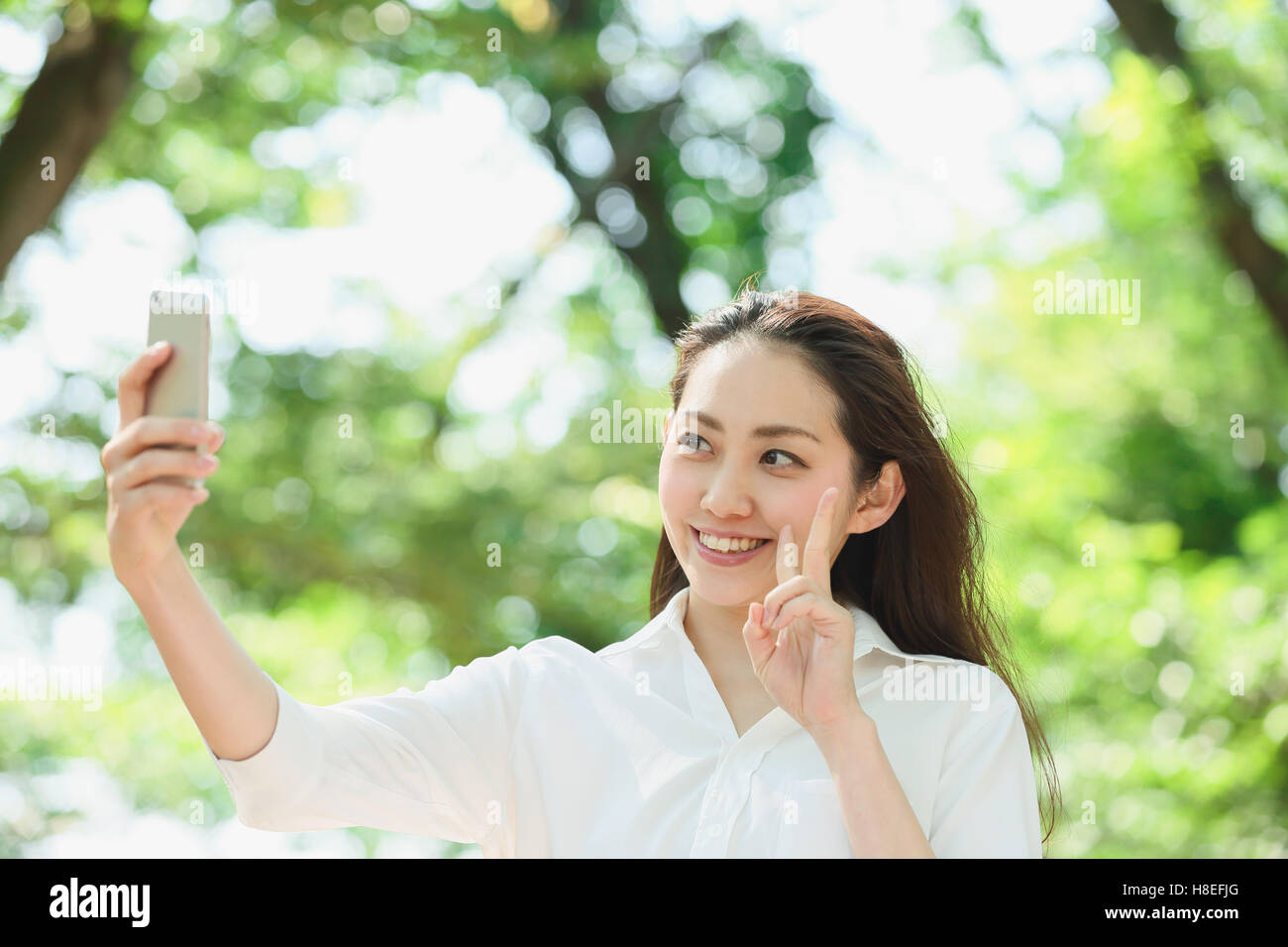 Young Japanese woman taking selfie surrounded by green in a city park Stock Photo