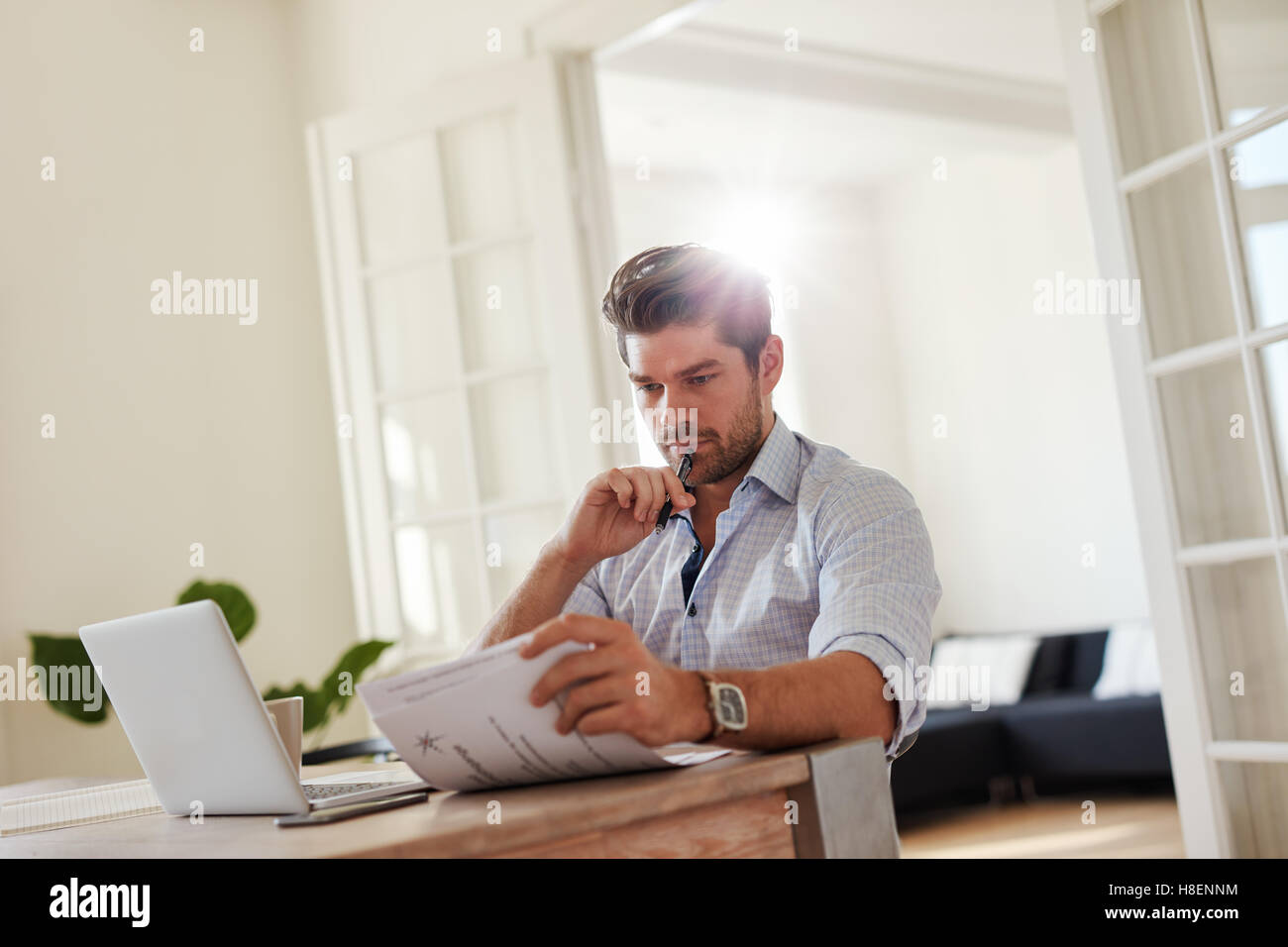 Shot of young man sitting at table with laptop and reading documents. Handsome business man working from home office. Stock Photo