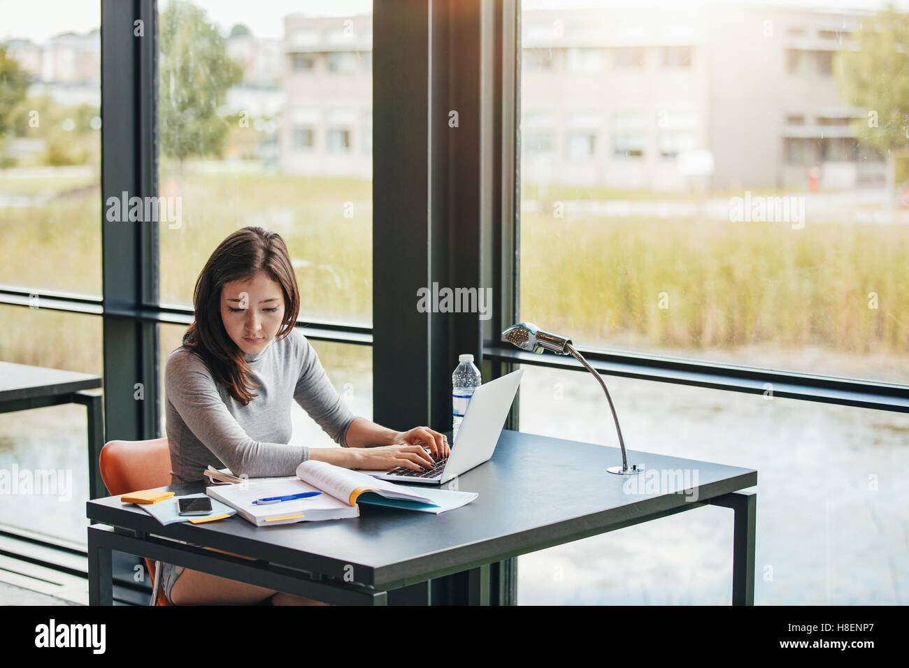 Young asian woman sitting in library searching information on laptop for her assignment. Young woman sitting at table reading bo Stock Photo