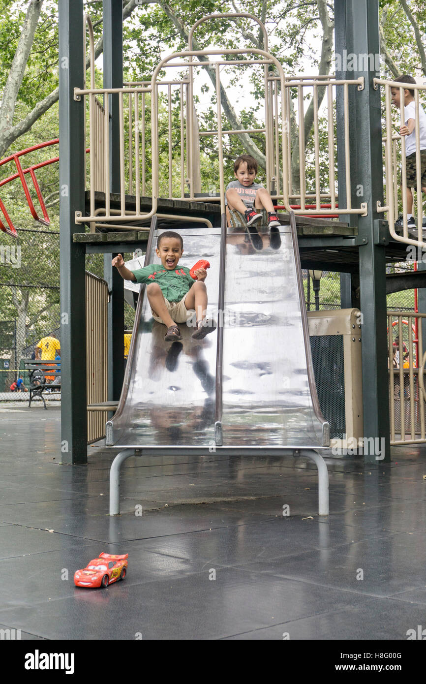 2 little boys play gleefully on playground slides with black child holding toy halfway on left & white child at top right slide Stock Photo