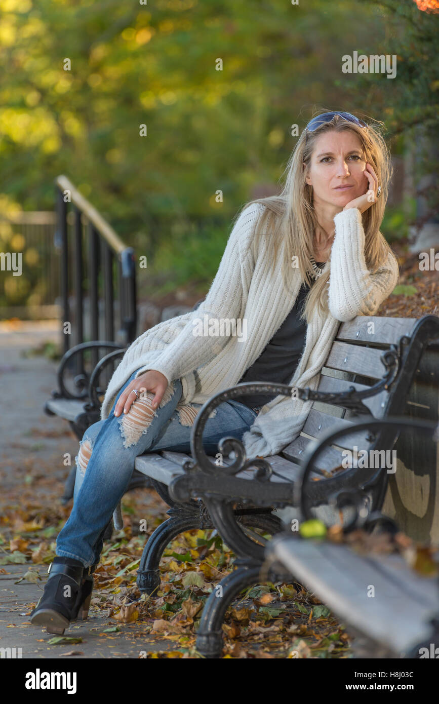 Attractive blond young woman sitting on bench in park with blurred background Stock Photo