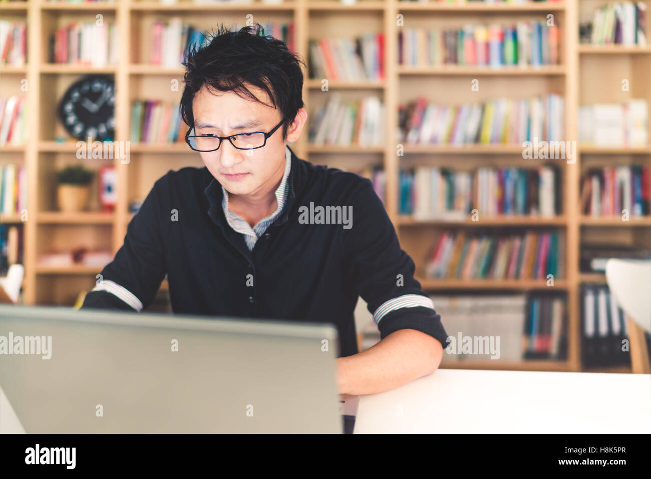 Young pensive Asian man working on laptop at home office or library with serious face, bookshelf blur background with copy space Stock Photo