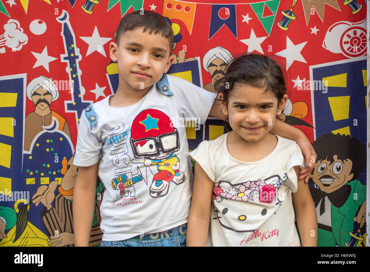 Cairo, Egypt. Young Egyptian boy and girl standing together in front of a colorful cartoon backdrop in the outdoor bazaar/ flea Stock Photo