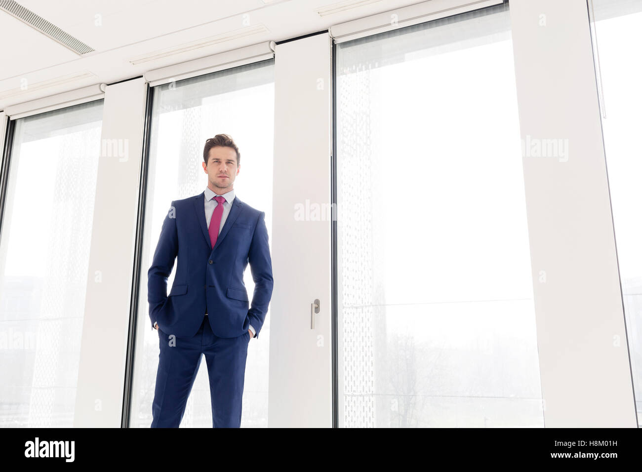 Portrait of confident young businessman standing with hands in pockets against office window Stock Photo