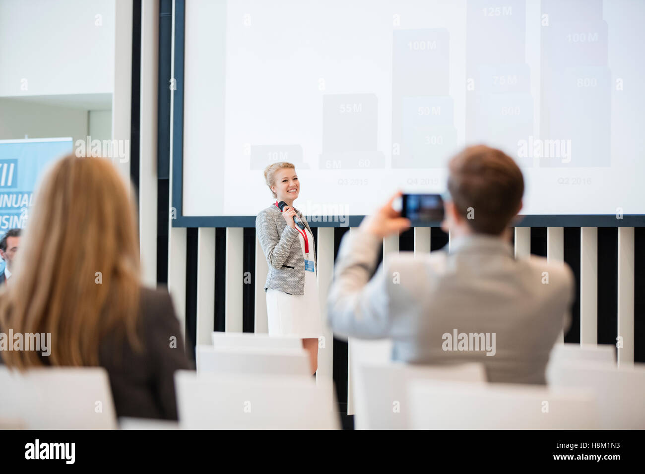 Rear view of businessman photographing female public speaker in seminar hall Stock Photo