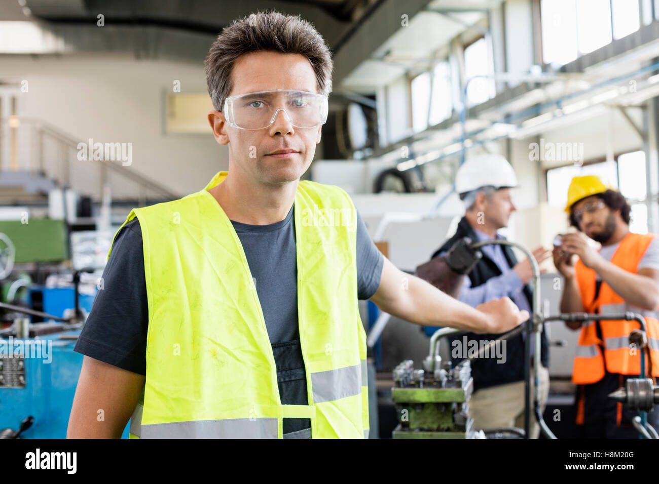 Portrait of mid adult worker wearing protective eyewear with colleagues in background at industry Stock Photo