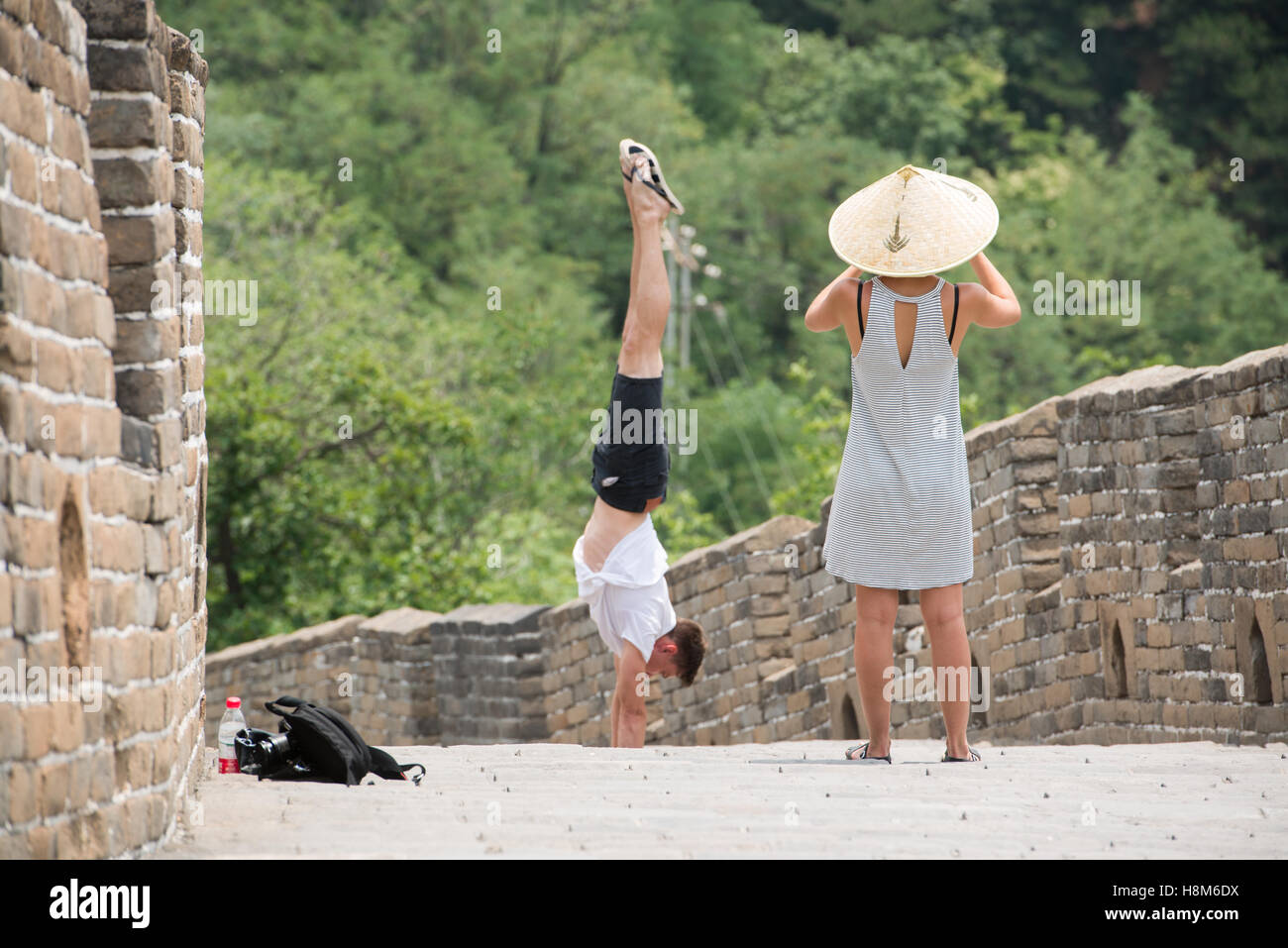 Mutianyu, China - Woman wearing an Asian conical hat taking pictures of a man doing a handstand on the Great Wall of China. The Stock Photo