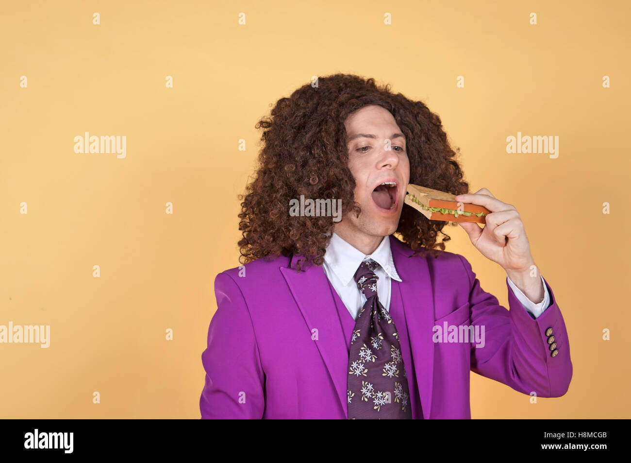 Caucasian man with afro wearing Purple Suit eating sandwich Stock Photo