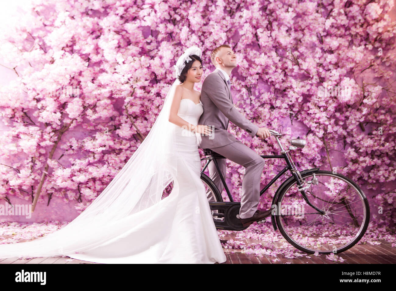 Thoughtful bride standing with bridegroom riding bicycle against pink flowers covering wall Stock Photo