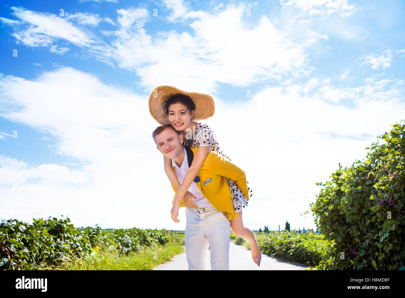 Portrait of happy man piggybacking woman on footpath amidst field against cloudy sky Stock Photo