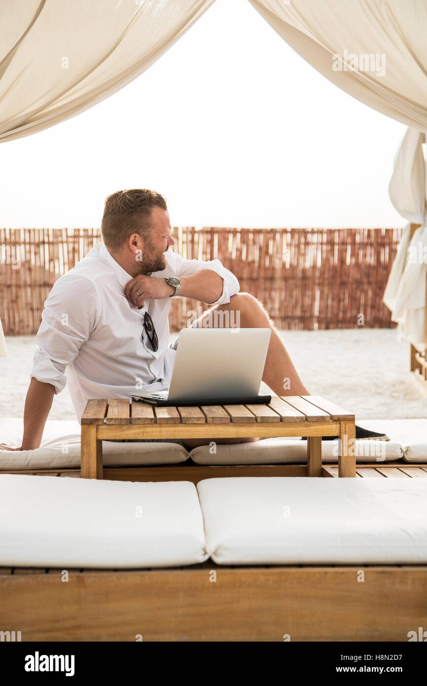 Mature man sitting on mattress on terrace under canopy and using laptop Stock Photo