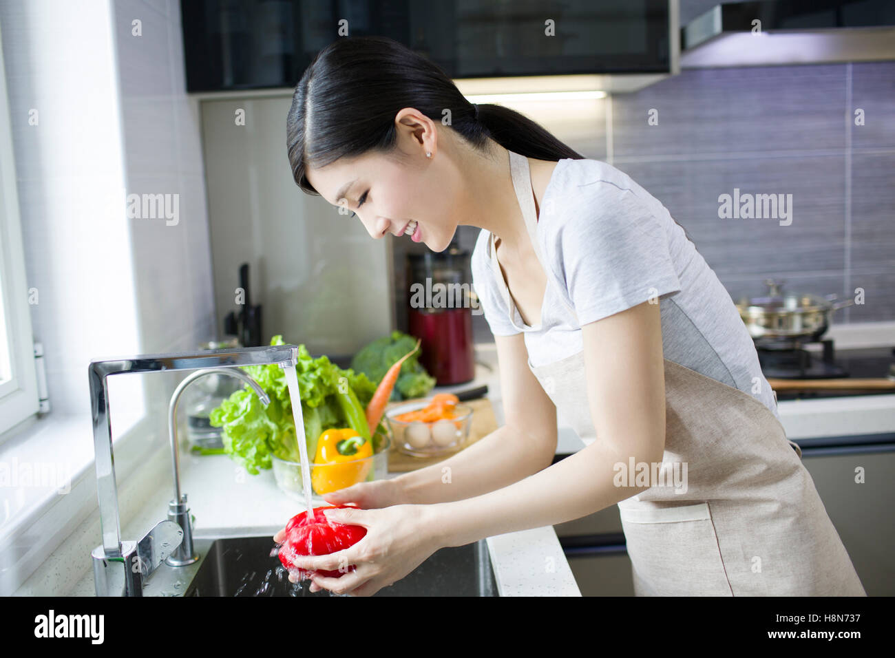 Happy young Chinese woman washing vegetables in kitchen Stock Photo