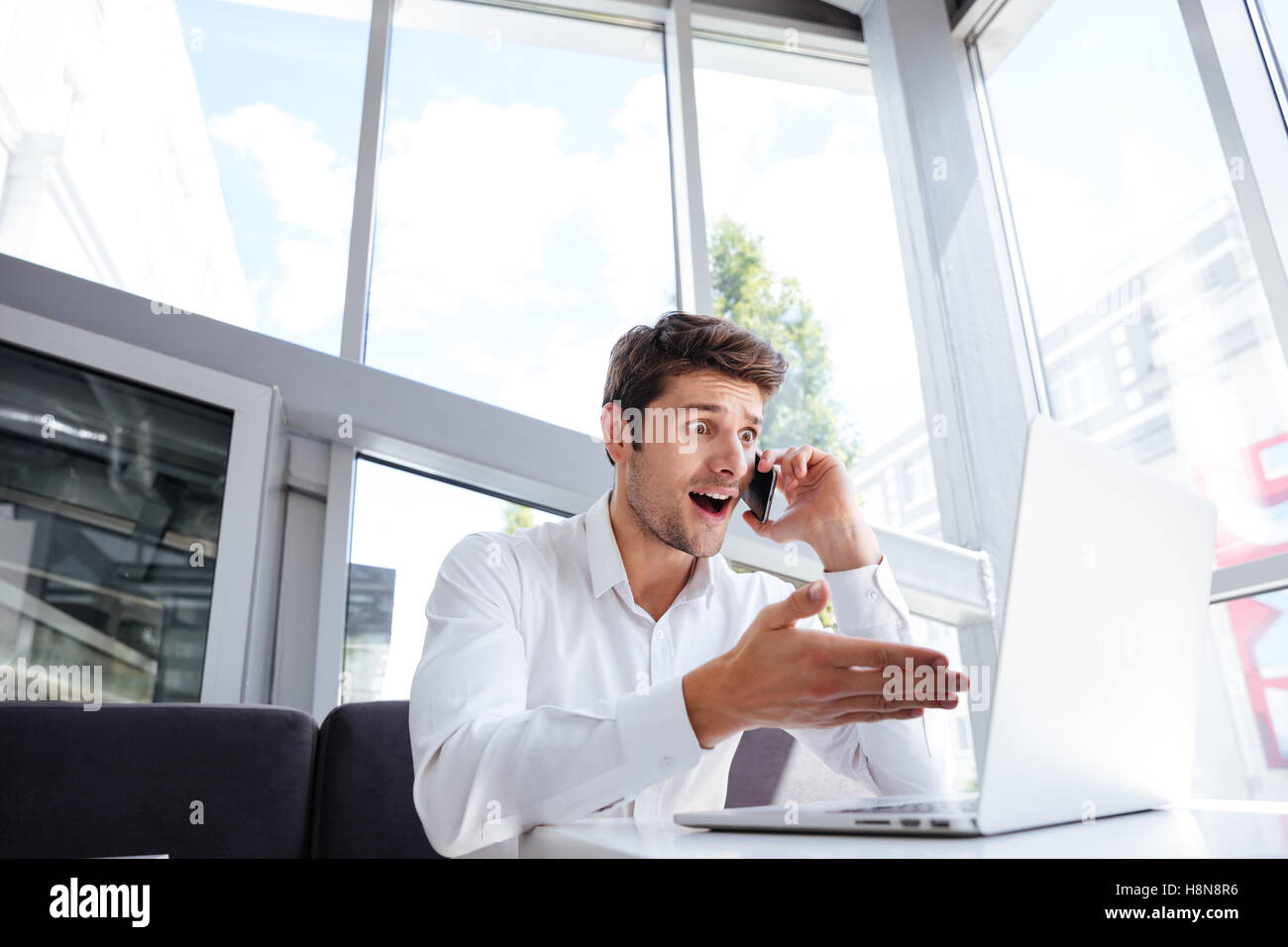 Amazed young businessman using laptop and talking on mobile phone in office Stock Photo