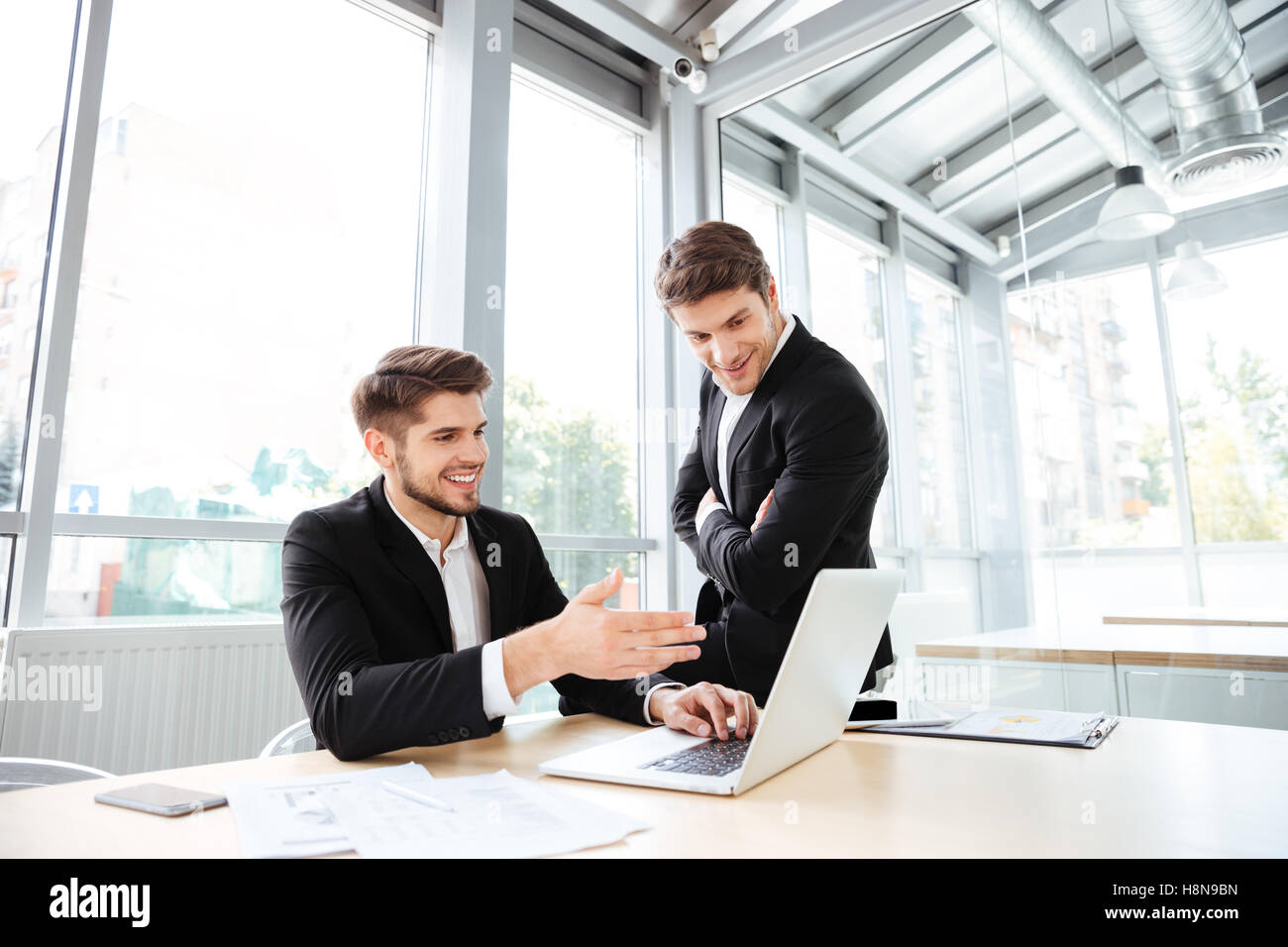 Two happy young businessmen talking and using laptop in office together ...