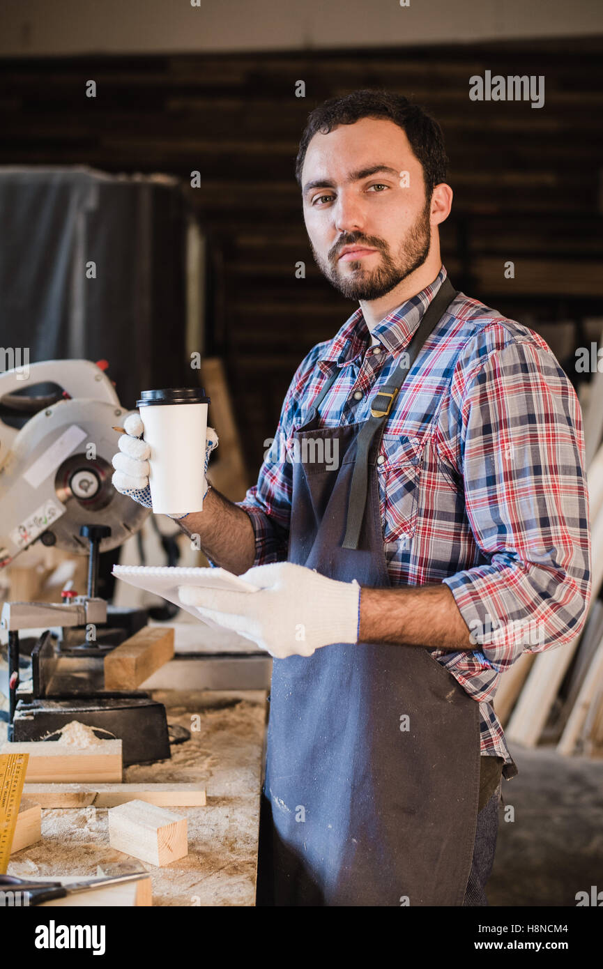 Carpenter taking a coffee break holding notebook in front of circular saw at his workshop Stock Photo